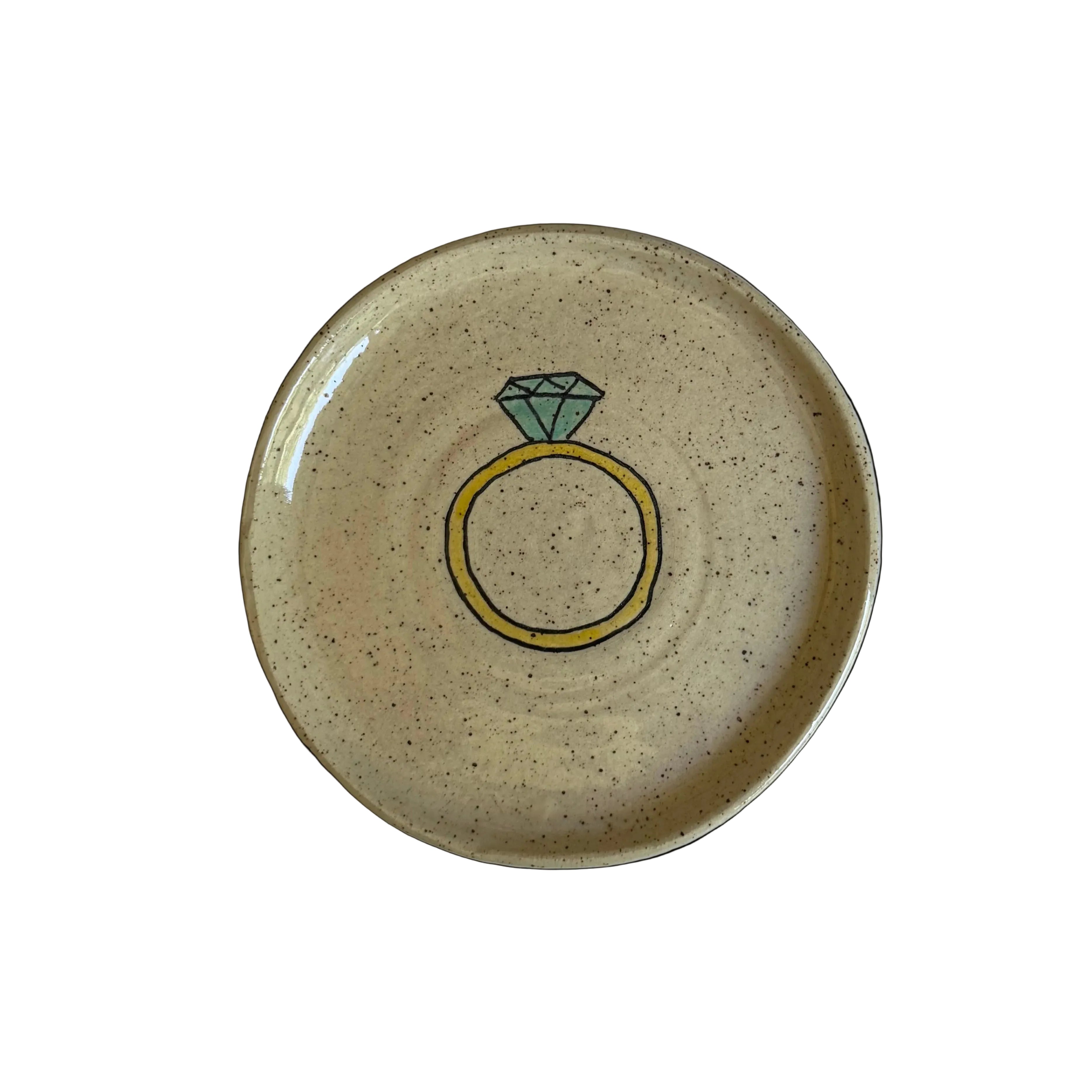 A round, speckled beige ceramic plate featuring a hand-drawn illustration of a yellow ring with a green diamond in the center, set against a plain light gray background.